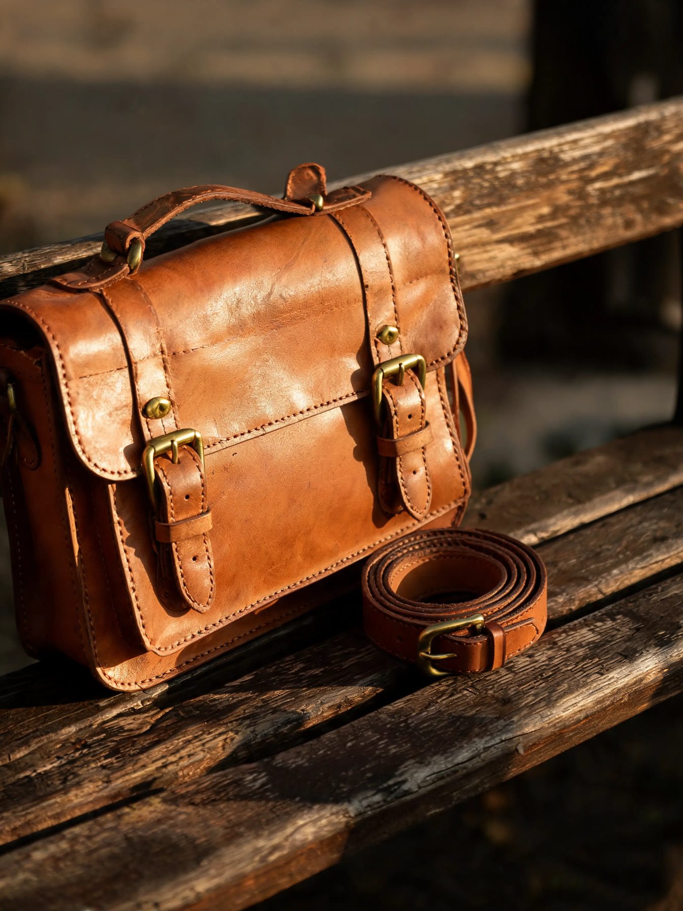 Vegetable-tanned leather satchel with brass buckles beside a coiled belt on a wooden bench